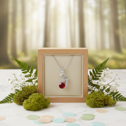 Silver necklace with a red gemstone in a wooden display box against a forest background