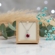 Silver necklace with a red gemstone pendant in a wooden box, surrounded by greenery and flowers.