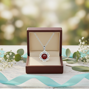 Silver necklace with a red pendant in an open jewelry box on a wooden surface with greenery.