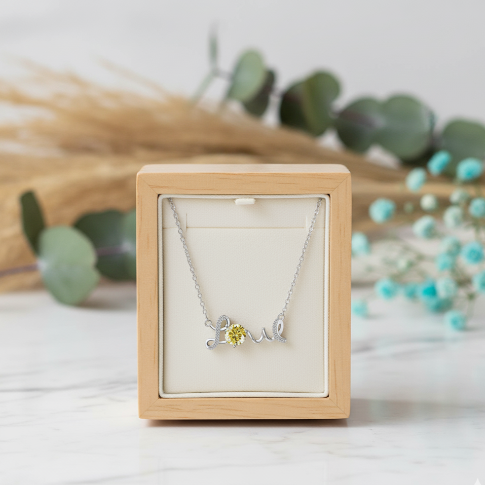 Silver necklace with a yellow center in a wooden box on a white surface with greenery.