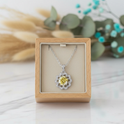 Silver necklace with a yellow center pendant in a wooden box on a light surface with greenery in the background