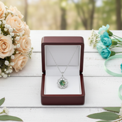 Silver necklace with green gemstone in a jewelry box on a wooden surface with flowers.