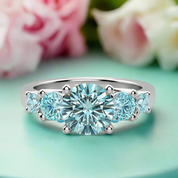 Silver ring with a central blue gemstone and smaller blue stones on a green surface with flowers in the background