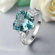 Silver ring with a heart-shaped blue gemstone and smaller clear stones on a light gray background.