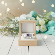 Silver ring with a purple gemstone in a wooden box on a wooden surface with flowers in the background.
