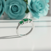 Silver ring with three green subject on a white table with tiffany colored flowers on display in the background with shadow on a white background
