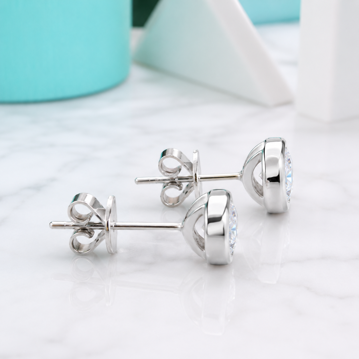 A pair of silver stud earrings with a small clear gemstone, displayed on a white marble surface with a soft-focus turquoise and white background.