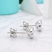 A pair of silver stud earrings with a small clear gemstone, displayed on a white marble surface with a soft-focus turquoise and white background.