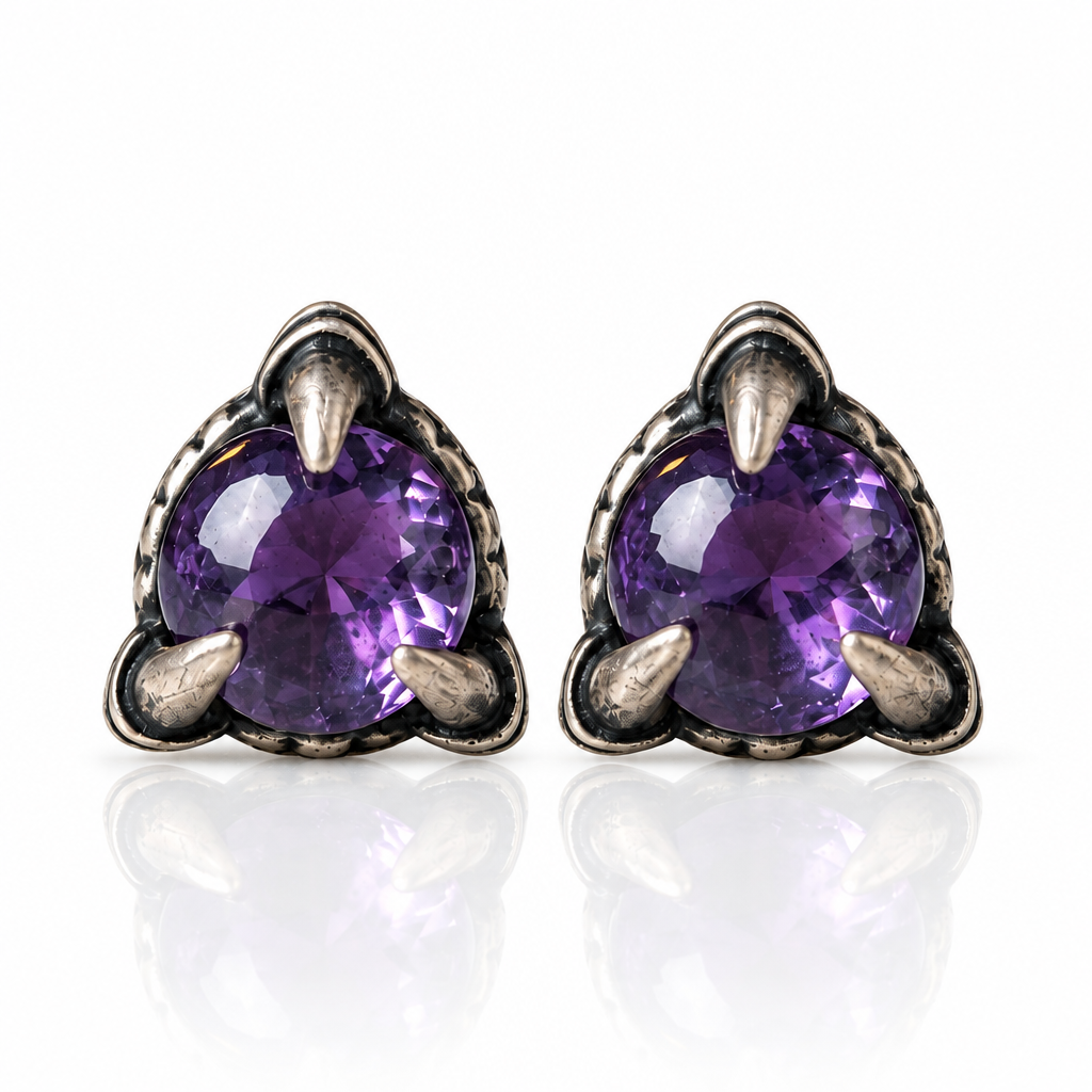 A pair of silver-toned stud earrings with oval-cut purple amethyst gemstones, displayed on a white marble surface with a lit green candle, red roses, and a green ribbon in the background.