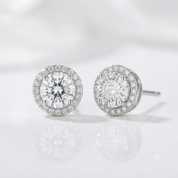 A pair of round halo stud earrings with brilliant-cut clear center stones and a surrounding halo of smaller clear stones in a silver-toned setting, displayed on a reflective white surface.