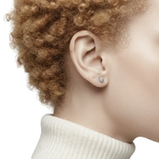 A close-up side profile of a person with curly light brown hair wearing a small round silver stud earring with a clear gemstone, paired with a cream ribbed turtleneck sweater against a light gray background.