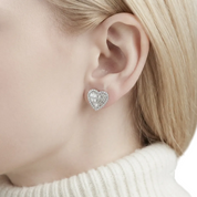A close-up of a woman's ear wearing a small heart-shaped stud earring with a silver-toned basketweave pattern of clear stones, paired with a cream ribbed turtleneck sweater.