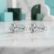 A pair of silver stud earrings with round-cut clear gemstones in a prong setting, displayed on a white marble surface with a soft-focus background.