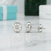 A pair of round bezel-set diamond stud earrings in silver-tone metal, shown on a white marble surface with a blurred turquoise gift box in the background.