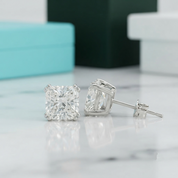 A pair of square princess-cut diamond stud earrings in a silver setting, with one earring facing forward and the other turned sideways to show the post and back. They are displayed on a white marble surface with a blurred turquoise box and a dark green box in the background.
