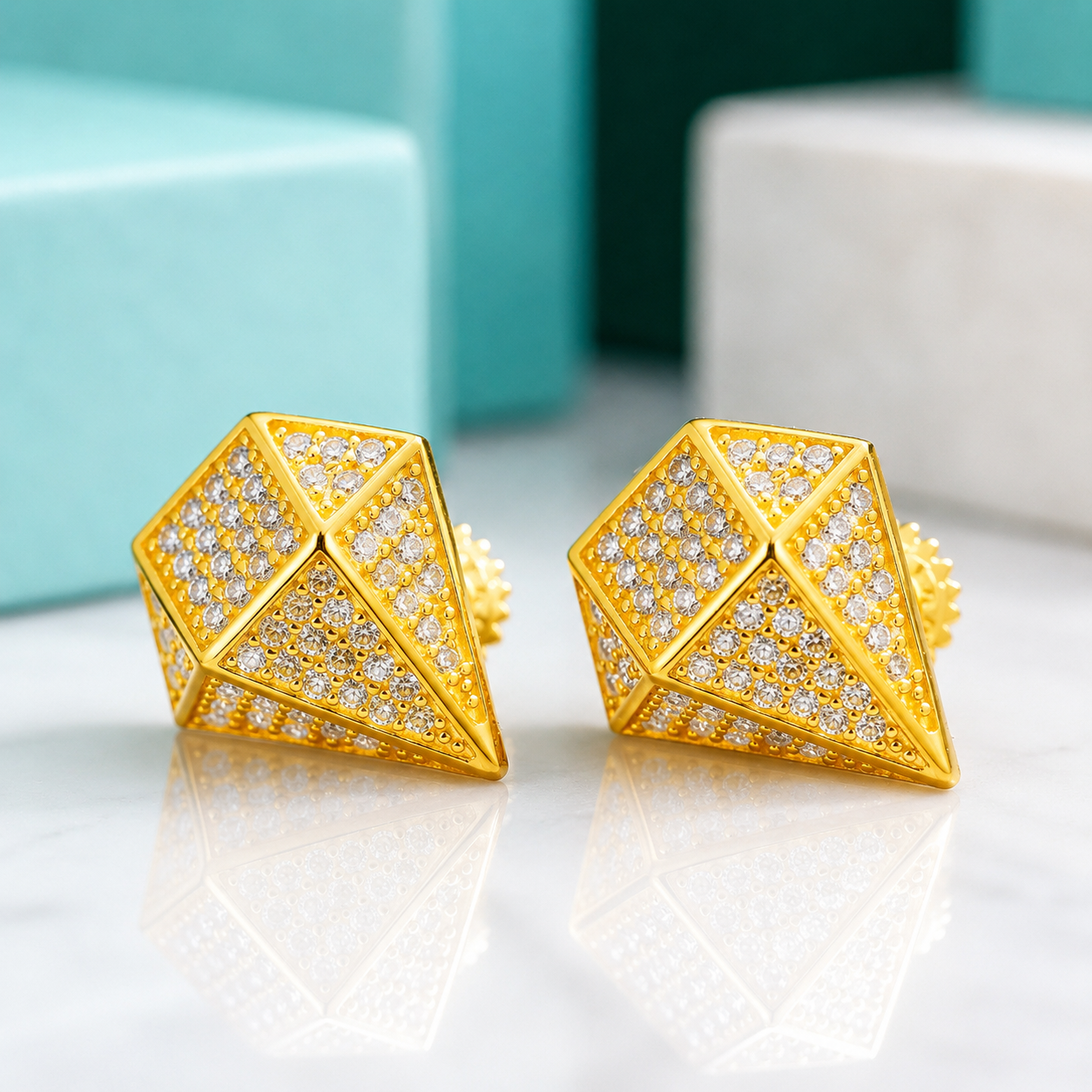 A pair of gold geometric stud earrings covered in small clear stones, displayed on a reflective white surface with blurred turquoise and white boxes in the background.