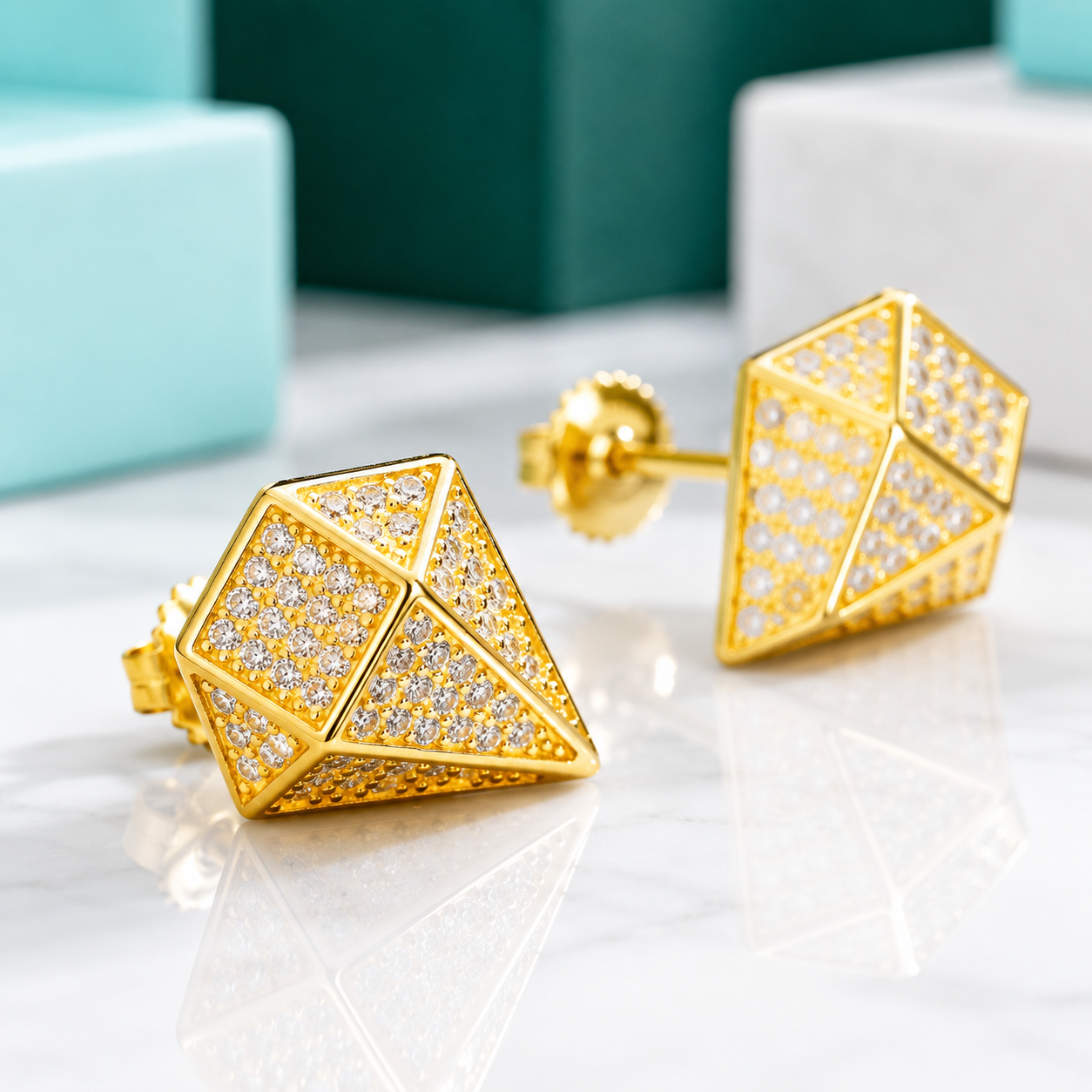 A pair of gold geometric stud earrings with pavé-set clear stones, displayed on a white marble surface with blurred turquoise and dark green gift boxes in the background.