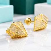 A pair of gold geometric stud earrings with pavé-set clear stones, displayed on a white marble surface with blurred turquoise and dark green gift boxes in the background.