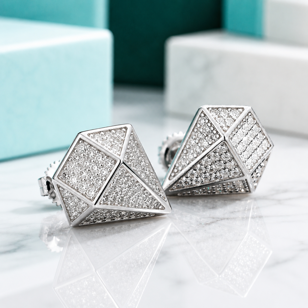 A pair of geometric stud earrings with a silver-toned frame and pavé-set clear stones, displayed on a white marble surface with blurred turquoise and white gift boxes in the background.