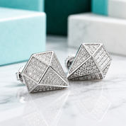 A pair of geometric stud earrings with a silver-toned frame and pavé-set clear stones, displayed on a white marble surface with blurred turquoise and white gift boxes in the background.