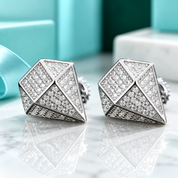 A pair of geometric stud earrings with a silver-toned diamond-cut design, heavily paved with clear cubic zirconia stones, displayed on a reflective white marble surface with a blurred turquoise jewelry box in the background.
