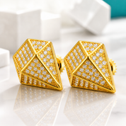 A pair of gold geometric stud earrings with pavé-set clear stones, displayed on a reflective white marble surface with blurred white blocks in the background.