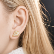 A close-up of a woman's ear wearing a small geometric gold stud earring with a textured, diamond-like pattern.