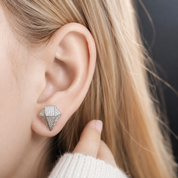 A close-up of a woman's ear wearing a small diamond-shaped stud earring set with clear stones in a silver-toned metal, with blonde hair and a white knit sweater visible.
