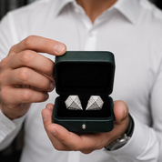 A close-up of a person in a white shirt holding an open dark green jewelry box. Inside the box are a pair of silver-toned geometric cufflinks with clear stones.