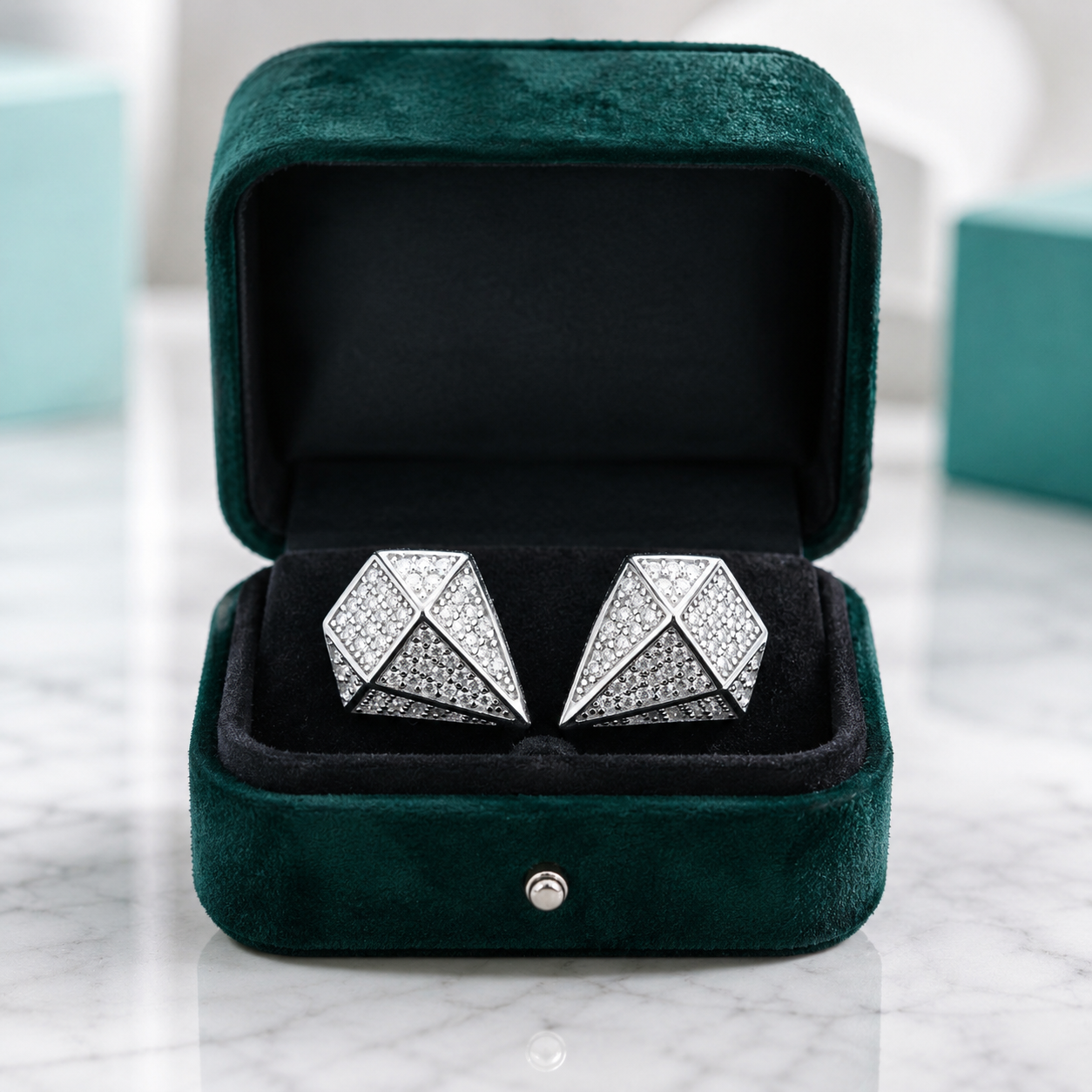 A pair of silver geometric stud earrings set with clear pavé stones, presented in an open dark green velvet jewelry box on a white marble surface.