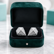 A pair of silver geometric stud earrings set with clear pavé stones, presented in an open dark green velvet jewelry box on a white marble surface.