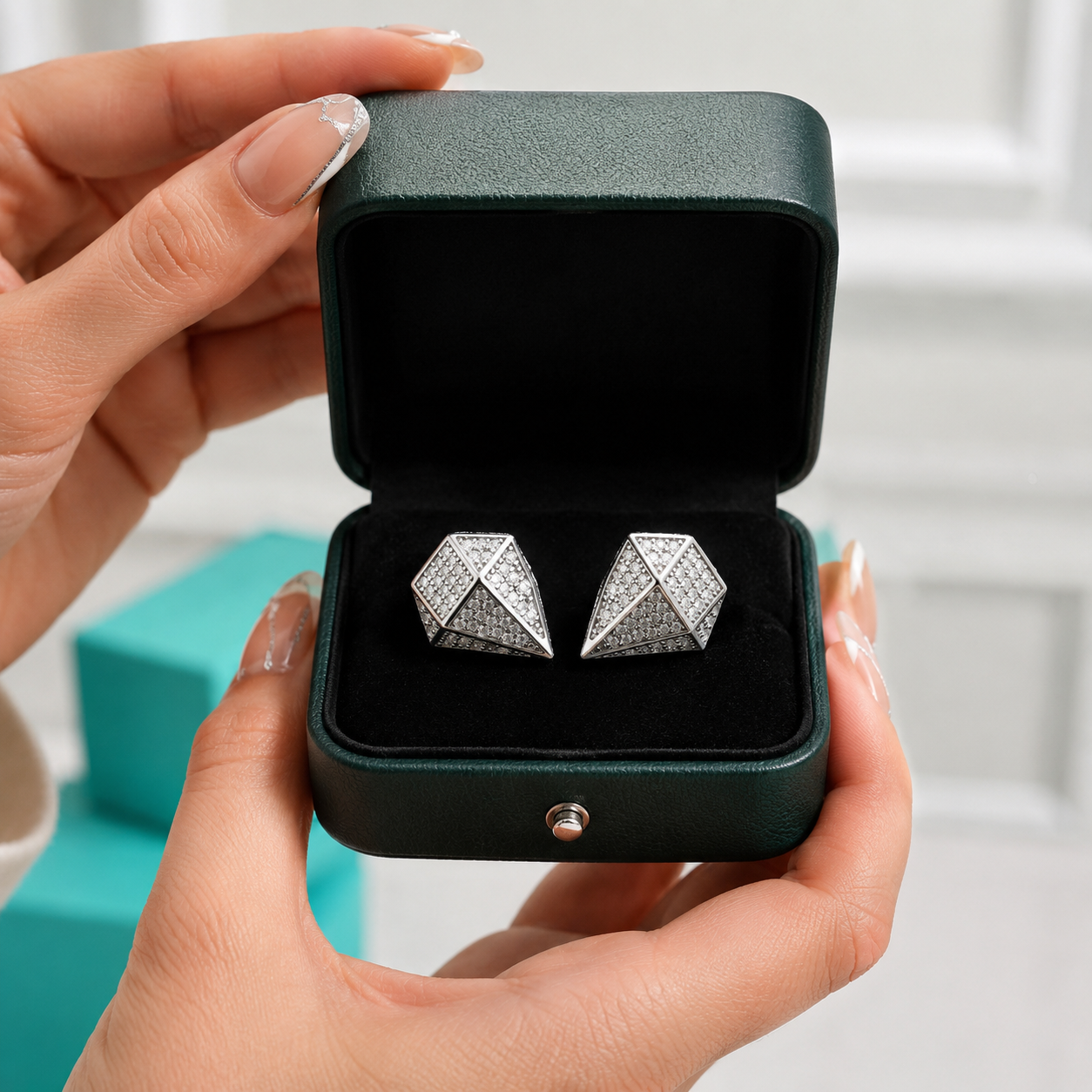 A pair of silver geometric stud earrings displayed in an open dark green jewelry box, held by hands with manicured nails.