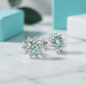 A pair of silver-toned snowflake-style stud earrings with round light aqua gemstones and surrounding clear stones, displayed on a white marble surface with blurred turquoise and white boxes in the background.
