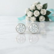 A pair of round bezel-set diamond stud earrings in a silver-toned metal, displayed on a white marble surface with a soft reflection. In the blurred background, a bouquet of white roses with a light blue ribbon is visible.