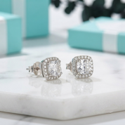 A pair of silver halo stud earrings with cushion-cut clear gemstones, displayed on a white marble surface with blurred turquoise gift boxes in the background.