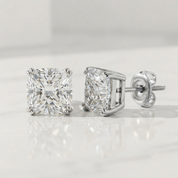A pair of princess-cut diamond stud earrings in a silver-colored four-prong setting, shown on a reflective white surface with a soft gray background.