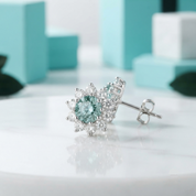 A pair of silver-toned stud earrings with a central round light blue gemstone surrounded by small clear stones, displayed on a white surface with soft turquoise blocks in the background.