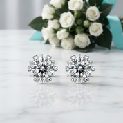A pair of silver-toned stud earrings with large clear faceted crystal designs, displayed on a white marble surface with white roses and a turquoise ribbon in the blurred background.