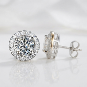 A pair of round halo stud earrings in silver-tone metal with clear gemstones, shown on a clean white reflective surface. One earring is facing forward to show the round cluster setting and post, while the other is angled to show the side profile of the back and post.