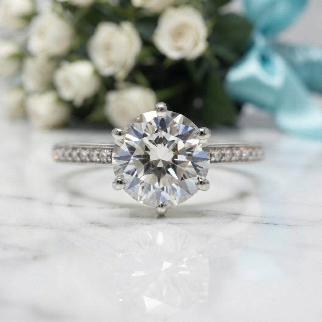 A silver solitaire engagement ring with a round brilliant-cut center stone and pavé-set side stones, displayed on a white marble surface with blurred white roses and a light blue ribbon in the background.
