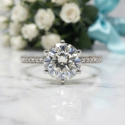 A silver solitaire engagement ring with a round brilliant-cut center stone and pavé-set side stones, displayed on a white marble surface with blurred white roses and a light blue ribbon in the background.