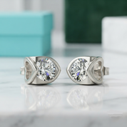 A pair of silver-toned stud earrings with large round clear gemstones, displayed on a light marble surface with blurred turquoise and green boxes in the background.