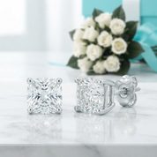 A pair of princess-cut solitaire stud earrings in silver-tone metal, displayed on a white marble surface with a bouquet of white roses and a turquoise gift box in the soft-focus background.
