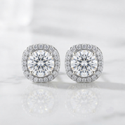A pair of round cushion-shaped diamond stud earrings with halo settings, displayed on a light marble surface with soft reflections.