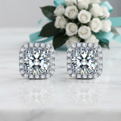 A pair of square cushion-cut halo stud earrings with brilliant round center stones and surrounding rows of small clear stones, displayed on a light marble surface with white roses and a turquoise ribbon in the background.