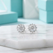 A pair of round silver stud earrings with clear gemstones, displayed on a white marble slab with blurred turquoise jewelry boxes in the background.