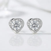 A pair of silver heart-shaped stud earrings with a large clear round center stone and a halo of smaller clear stones, displayed on a clean white reflective surface.