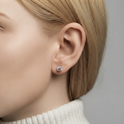 A close-up of a woman's ear wearing a small round silver stud earring with a clear gemstone, set against a neutral gray background.
