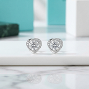 A pair of heart-shaped stud earrings with clear gemstones in a silver-toned setting, displayed on a white marble surface with blurred turquoise and white boxes in the background.