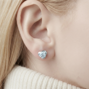 A close-up of a person's ear wearing a small heart-shaped stud earring with a silver-toned setting and clear stones.